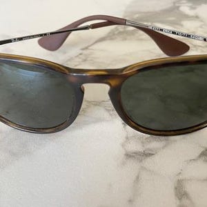 K&ouml;nnte beinhalten: Sonnenbrille mit Schildpatt-Fassung und dunklen Gl&auml;sern. Die Sonnenbrille hat eine klassische Cat-Eye-Form mit einem braun-schwarzen Muster auf den Fassungen. Die B&uuml;gel sind dunkelbraun und die Gl&auml;ser sind dunkelgr&uuml;n.