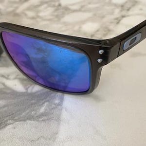 May include: Dark brown sunglasses with blue and purple reflective lenses. The sunglasses have a rectangular frame and a small logo on the temple. The lenses are slightly curved.