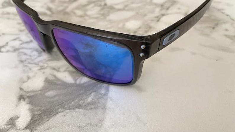 May include: Dark brown sunglasses with blue and purple reflective lenses. The sunglasses have a rectangular frame and a small logo on the temple. The lenses are slightly curved.