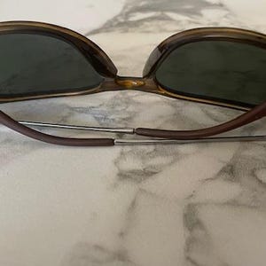 K&ouml;nnte beinhalten: Sonnenbrille mit braunem Schildpattrahmen und dunkelgr&uuml;nen Gl&auml;sern. Die B&uuml;gel sind braun und mit silbernen Scharnieren am Rahmen befestigt. Die Sonnenbrille liegt auf einer wei&szlig;-grauen Marmoroberfl&auml;che.