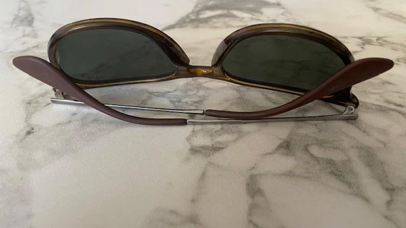 K&ouml;nnte beinhalten: Sonnenbrille mit braunem Schildpattrahmen und dunkelgr&uuml;nen Gl&auml;sern. Die B&uuml;gel sind braun und mit silbernen Scharnieren am Rahmen befestigt. Die Sonnenbrille liegt auf einer wei&szlig;-grauen Marmoroberfl&auml;che.