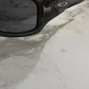 May include: Close-up of a pair of dark gray sunglasses with a black frame. The sunglasses have a curved design and a small silver logo on the temple. The lenses are dark and reflective.