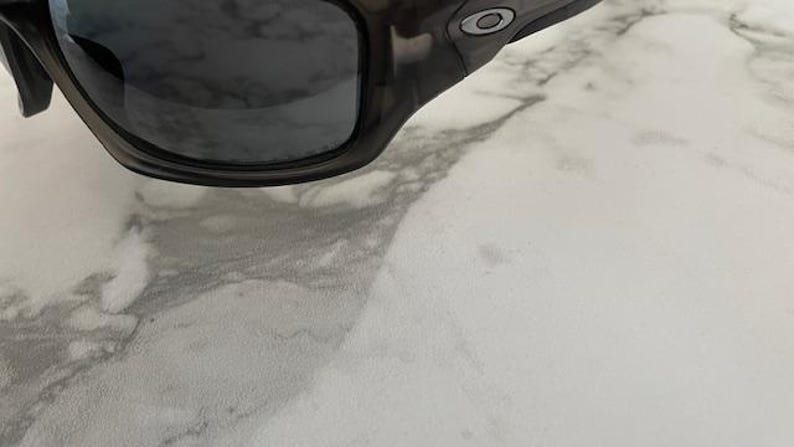 May include: Close-up of a pair of dark gray sunglasses with a black frame. The sunglasses have a curved design and a small silver logo on the temple. The lenses are dark and reflective.