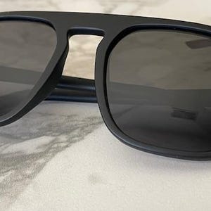 Puede incluir: Gafas de sol con montura negra mate y lentes oscuras. El dise&ntilde;o presenta una forma de lente cuadrada con una barra superior prominente que conecta ambos aros, creando una l&iacute;nea de ceja distintiva con varillas delgadas que se extienden hacia afuera.