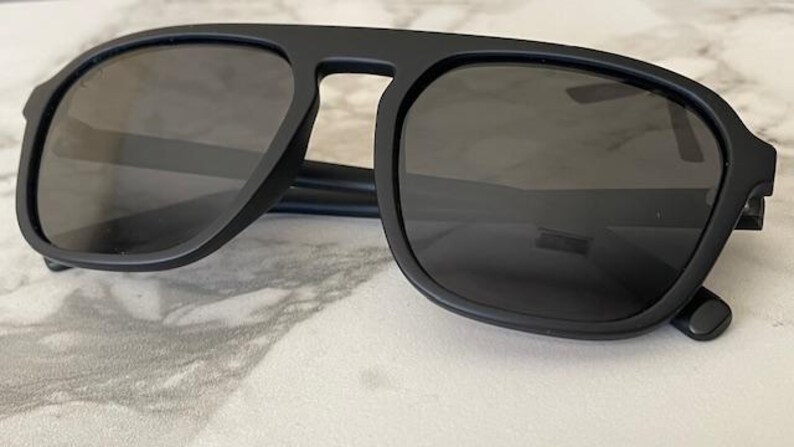 Puede incluir: Gafas de sol con montura negra mate y lentes oscuras. El dise&ntilde;o presenta una forma de lente cuadrada con una barra superior prominente que conecta ambos aros, creando una l&iacute;nea de ceja distintiva con varillas delgadas que se extienden hacia afuera.