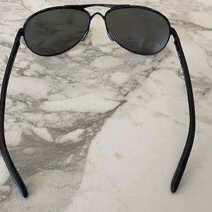 May include: Black aviator sunglasses with dark lenses. The sunglasses have a double bridge and a thin, black metal frame. The temples curve gently, and the sunglasses are set against a marble background.