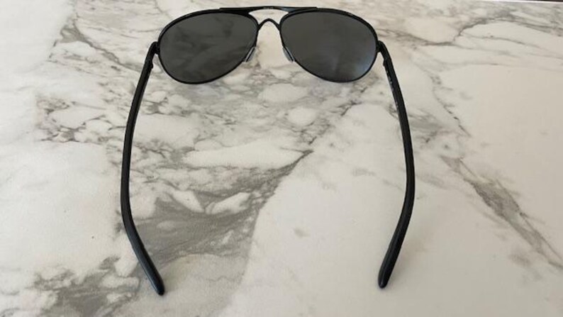 May include: Black aviator sunglasses with dark lenses. The sunglasses have a double bridge and a thin, black metal frame. The temples curve gently, and the sunglasses are set against a marble background.