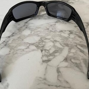 K&ouml;nnte beinhalten: Schwarze Sonnenbrille mit dunkelgrauen Gl&auml;sern. Die Sonnenbrille hat ein sportliches Design mit einem dicken Rahmen und gebogenen B&uuml;geln. Die Gl&auml;ser sind rechteckig und der Rahmen besteht aus gl&auml;nzendem Material.