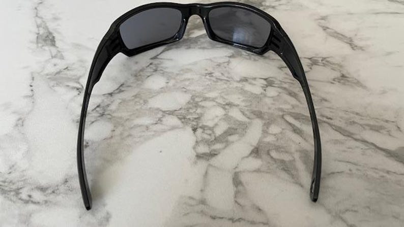 K&ouml;nnte beinhalten: Schwarze Sonnenbrille mit dunkelgrauen Gl&auml;sern. Die Sonnenbrille hat ein sportliches Design mit einem dicken Rahmen und gebogenen B&uuml;geln. Die Gl&auml;ser sind rechteckig und der Rahmen besteht aus gl&auml;nzendem Material.