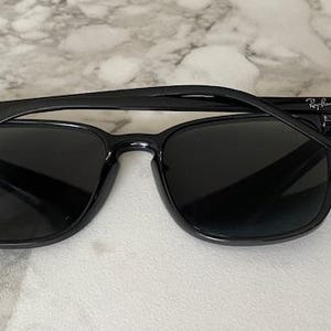 Puede incluir: Gafas de sol negras con montura cuadrada. Las gafas tienen lentes oscuras y una montura negra brillante. El logotipo de Ray-Ban es visible en la patilla. Las gafas descansan sobre una superficie de m&aacute;rmol.