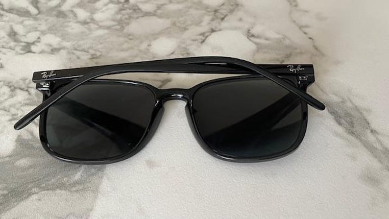 Puede incluir: Gafas de sol negras con montura cuadrada. Las gafas tienen lentes oscuras y una montura negra brillante. El logotipo de Ray-Ban es visible en la patilla. Las gafas descansan sobre una superficie de m&aacute;rmol.