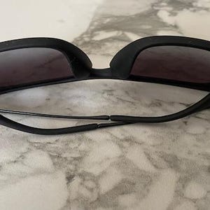 Puede incluir: Gafas de sol negras con lentes oscuras. Las gafas tienen una montura y patillas negras mate gruesas. Las lentes son de color gris oscuro y las gafas descansan sobre una superficie clara.