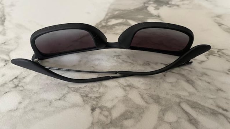 Puede incluir: Gafas de sol negras con lentes oscuras. Las gafas tienen una montura y patillas negras mate gruesas. Las lentes son de color gris oscuro y las gafas descansan sobre una superficie clara.