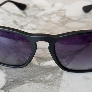 May include: Black sunglasses with dark purple lenses. The sunglasses have a matte black frame and thin black arms. The Ray-Ban logo is visible on the left lens. The sunglasses are resting on a light-colored surface.