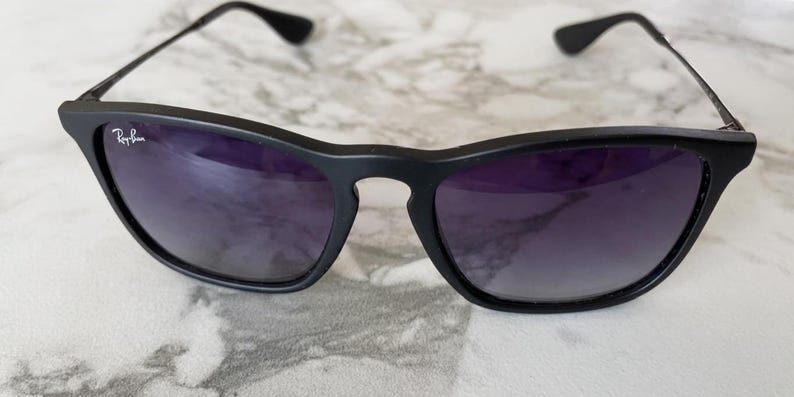 May include: Black sunglasses with dark purple lenses. The sunglasses have a matte black frame and thin black arms. The Ray-Ban logo is visible on the left lens. The sunglasses are resting on a light-colored surface.