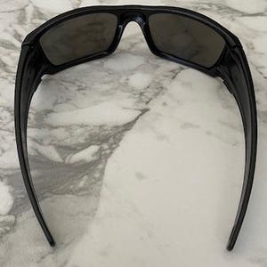 May include: Black sunglasses with a rectangular frame and dark lenses. The sunglasses have a sleek, modern design with curved arms. The image is taken from above, showing the sunglasses against a marble background.