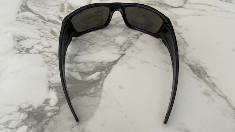May include: Black sunglasses with a rectangular frame and dark lenses. The sunglasses have a sleek, modern design with curved arms. The image is taken from above, showing the sunglasses against a marble background.