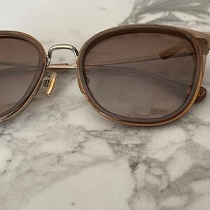May include: Brown-tinted sunglasses with a rounded frame and gold-colored metal accents. The lenses are a gradient brown color. The sunglasses are resting on a white and gray marble surface.