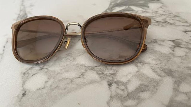 May include: Brown-tinted sunglasses with a rounded frame and gold-colored metal accents. The lenses are a gradient brown color. The sunglasses are resting on a white and gray marble surface.