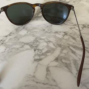 K&ouml;nnte beinhalten: Dunkelbraune Sonnenbrille mit rundem Rahmen und dunklen Gl&auml;sern. Die B&uuml;gel sind aus d&uuml;nnem Metall mit braunen Spitzen. Die Sonnenbrille liegt auf einem Marmorhintergrund.