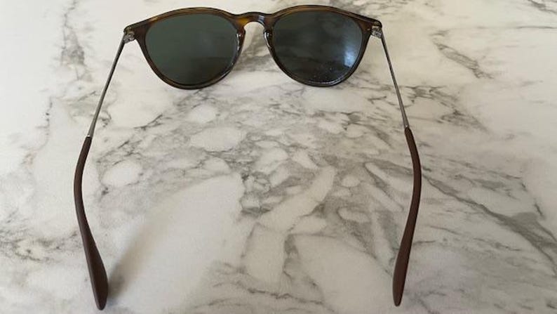 K&ouml;nnte beinhalten: Dunkelbraune Sonnenbrille mit rundem Rahmen und dunklen Gl&auml;sern. Die B&uuml;gel sind aus d&uuml;nnem Metall mit braunen Spitzen. Die Sonnenbrille liegt auf einem Marmorhintergrund.