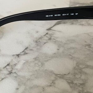 Puede incluir: Gafas de sol negras con montura rectangular y lentes verdes. La patilla lleva el texto "RB 4194 601/8A 53口17 140 3P". Las gafas de sol est&aacute;n sobre un fondo de m&aacute;rmol.