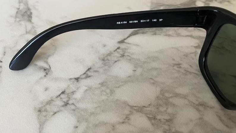 Puede incluir: Gafas de sol negras con montura rectangular y lentes verdes. La patilla lleva el texto "RB 4194 601/8A 53口17 140 3P". Las gafas de sol est&aacute;n sobre un fondo de m&aacute;rmol.