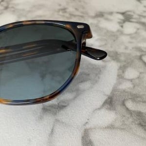 May include: Sunglasses with a tortoiseshell pattern frame and blue-tinted lenses. The frame features a mix of brown, orange, and dark blue hues. The sunglasses have a classic, rounded shape, suitable for various face shapes.