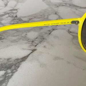 May include: Bright yellow sunglasses with dark lenses. The frame is a vibrant yellow, and the lenses are a dark, neutral color. The sunglasses have a classic, rounded shape. The text "RB4125 CATS 5000 764/32 IN" is printed on the arm.