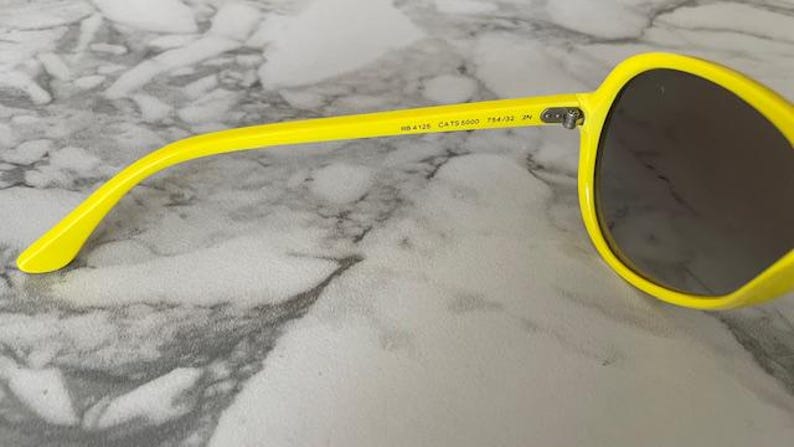 May include: Bright yellow sunglasses with dark lenses. The frame is a vibrant yellow, and the lenses are a dark, neutral color. The sunglasses have a classic, rounded shape. The text "RB4125 CATS 5000 764/32 IN" is printed on the arm.