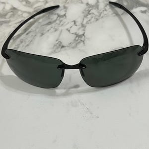 May include: Rimless sunglasses with dark lenses and black frames. The sunglasses have a sleek, modern design, with a bridge connecting the two lenses. The temples curve gently, providing a comfortable fit. The sunglasses are suitable for outdoor activities.
