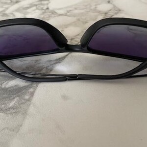 May include: Black sunglasses with purple-tinted lenses. The sunglasses have a sleek, modern design with a matte finish. The arms are thin and connect to the frame with a hinge. The sunglasses are resting on a light-colored surface.