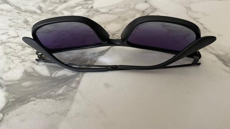May include: Black sunglasses with purple-tinted lenses. The sunglasses have a sleek, modern design with a matte finish. The arms are thin and connect to the frame with a hinge. The sunglasses are resting on a light-colored surface.