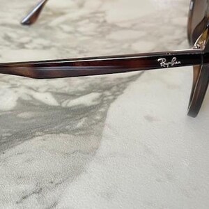 May include: Brown tortoiseshell sunglasses with a dark frame and lenses. The Ray-Ban logo is visible on the temple. The sunglasses are resting on a light-colored, patterned surface.