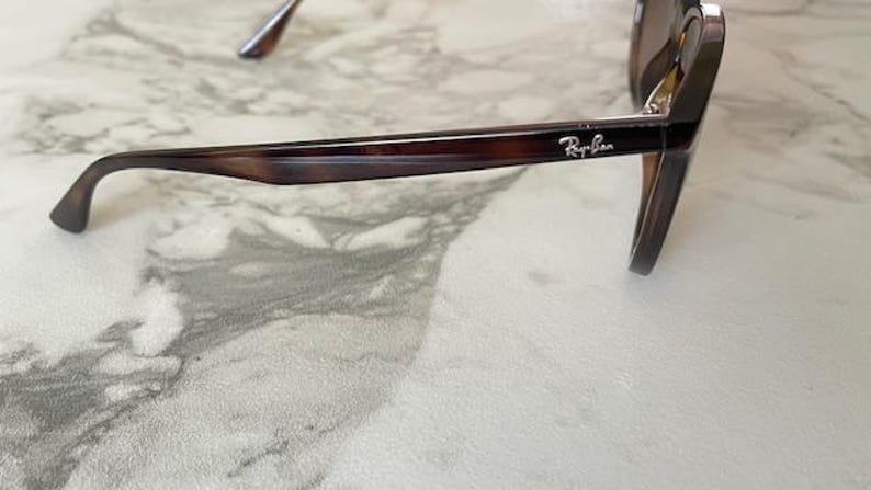 May include: Brown tortoiseshell sunglasses with a dark frame and lenses. The Ray-Ban logo is visible on the temple. The sunglasses are resting on a light-colored, patterned surface.
