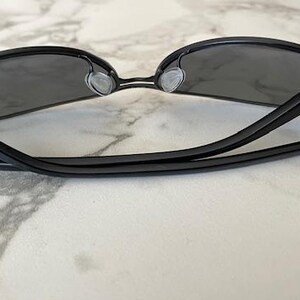 May include: Black sunglasses with dark gray lenses. The sunglasses have a sleek, modern design with a thin frame and a bridge over the nose. The temples are straight and extend back to the ear pieces.