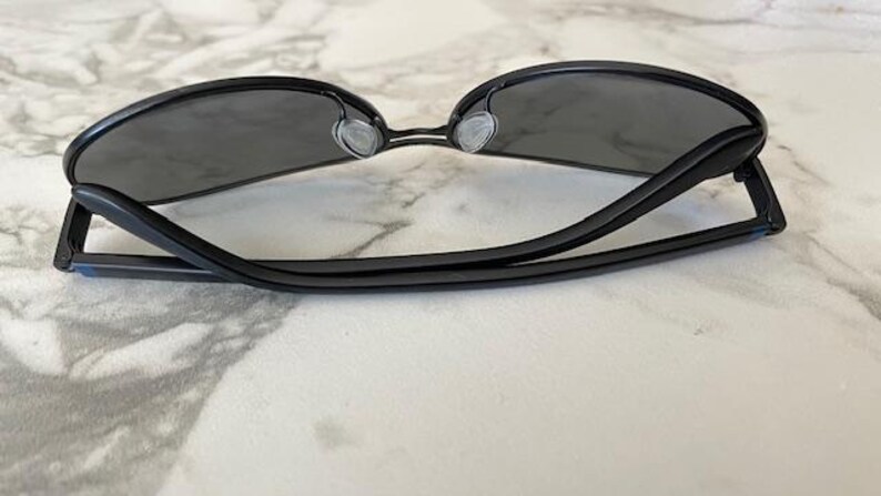 May include: Black sunglasses with dark gray lenses. The sunglasses have a sleek, modern design with a thin frame and a bridge over the nose. The temples are straight and extend back to the ear pieces.