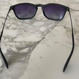 May include: Black sunglasses with a square frame and gradient lenses. The sunglasses have a sleek, modern design with thin temples. The lenses transition from a darker shade at the top to a lighter shade at the bottom.