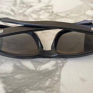 May include: Black sunglasses with dark lenses, folded on a marble surface. The sunglasses have a rectangular frame with a silver accent on the temples. The arms are folded over the lenses.