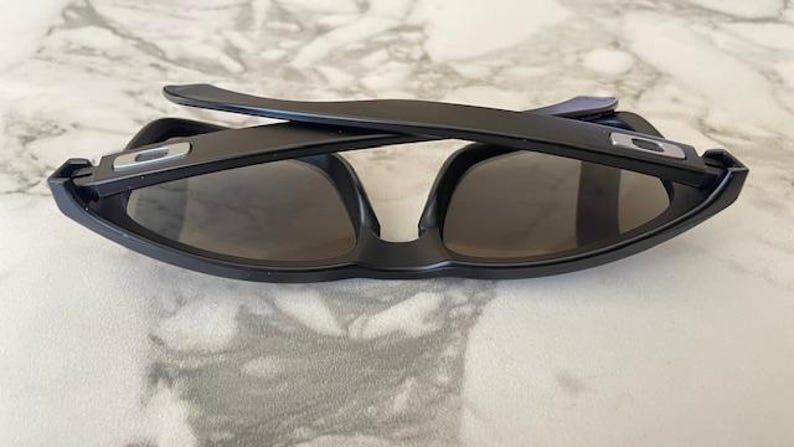 May include: Black sunglasses with dark lenses, folded on a marble surface. The sunglasses have a rectangular frame with a silver accent on the temples. The arms are folded over the lenses.