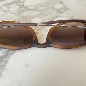 May include: Brown sunglasses with a unique frame design. The lenses are a gradient brown color, and the arms are a darker brown. The sunglasses have a silver metal bridge and hinges. The glasses are resting on a white and gray surface.