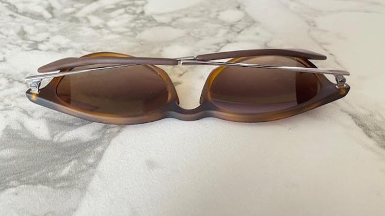 May include: Brown sunglasses with a unique frame design. The lenses are a gradient brown color, and the arms are a darker brown. The sunglasses have a silver metal bridge and hinges. The glasses are resting on a white and gray surface.