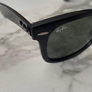 K&ouml;nnte beinhalten: Schwarze Sonnenbrille mit dunklen Gl&auml;sern. Das Ray-Ban-Logo ist auf dem Glas sichtbar. Die Sonnenbrille hat ein klassisches, rechteckiges Rahmendesign. Die Scharniere und B&uuml;gel sind ebenfalls schwarz. Auf einer Marmoroberfl&auml;che.