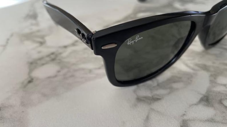 K&ouml;nnte beinhalten: Schwarze Sonnenbrille mit dunklen Gl&auml;sern. Das Ray-Ban-Logo ist auf dem Glas sichtbar. Die Sonnenbrille hat ein klassisches, rechteckiges Rahmendesign. Die Scharniere und B&uuml;gel sind ebenfalls schwarz. Auf einer Marmoroberfl&auml;che.