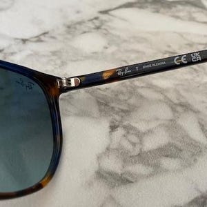 May include: Sunglasses with blue lenses and a tortoiseshell pattern on the frames. The temples have a dark blue and brown pattern. The Ray-Ban logo is visible on the temple and lens.