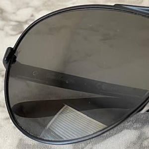 May include: Close-up of black aviator sunglasses with dark gray lenses. The sunglasses have a sleek, modern design with a thin frame and a bridge. The lenses are slightly reflective.