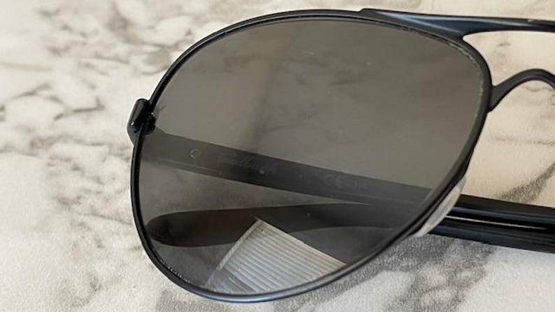 May include: Close-up of black aviator sunglasses with dark gray lenses. The sunglasses have a sleek, modern design with a thin frame and a bridge. The lenses are slightly reflective.