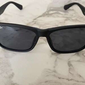 Pu&ograve; includere: Occhiali da sole neri con lenti scure. Gli occhiali hanno una montatura rettangolare con bordi arrotondati e una finitura opaca. Il logo Ray-Ban &egrave; visibile sulla lente sinistra. Gli occhiali sono appoggiati su una superficie di marmo.