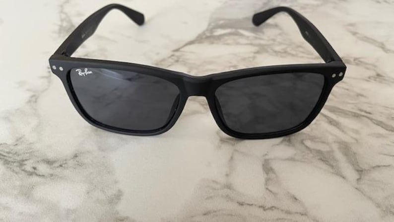 Pu&ograve; includere: Occhiali da sole neri con lenti scure. Gli occhiali hanno una montatura rettangolare con bordi arrotondati e una finitura opaca. Il logo Ray-Ban &egrave; visibile sulla lente sinistra. Gli occhiali sono appoggiati su una superficie di marmo.