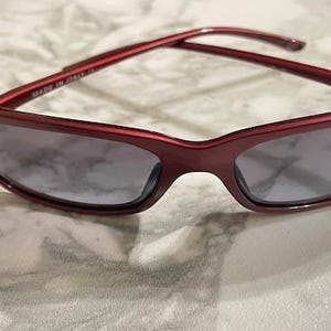 May include: Red-framed sunglasses with gray lenses. The sunglasses have a rectangular shape and a glossy finish. The image shows the sunglasses on a marble-patterned surface. The text "MADE IN ITALY" is visible on the frame.